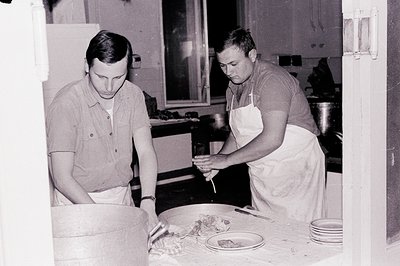 Two men in short-sleeved shirts and aprons are shown in a workspace, likely preparing food. One kneads dough in a large woode...