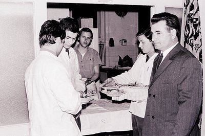A formal gathering; a man in a suit observes two people in lab coats presenting food on trays in a tiled kitchen. Possibly a ...