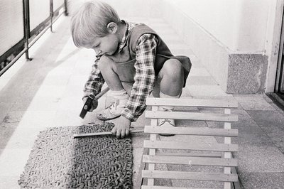 A young boy, crouched on a textured mat, appears engrossed in using a small hammer and tool on a wooden structure. He's weari...