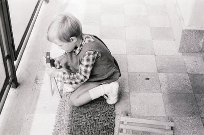A young boy, possibly 6-8 years old, kneels near a building’s glass entrance, intently focused on a small, boxy camera. He we...