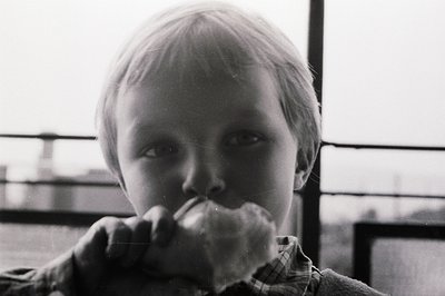 Close-up, black and white photo captures a young boy with short blond hair, intently eating what appears to be a large sandwi...
