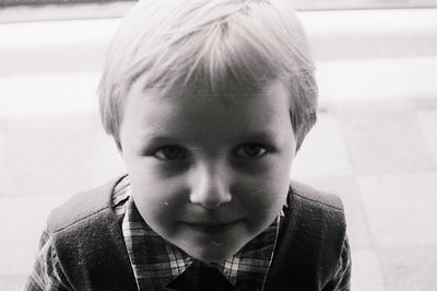 Close-up, candid portrait of a young boy with short, light hair. He wears a plaid shirt and jacket, looking directly at the c...
