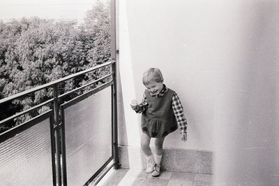 A young boy in a checked shirt and bloomers playfully poses on a balcony. The black and white image likely dates to the 1960s...