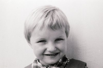 Close-up portrait of a young boy with short, blond hair and a cheerful grin. He's wearing a plaid shirt and a jacket. Plain b...