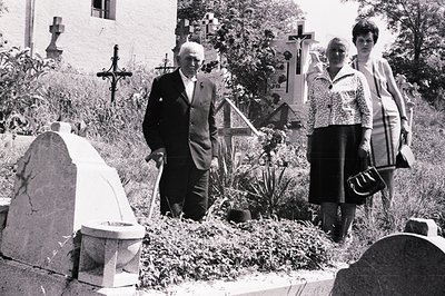 Monochrome image of an elderly man in a suit and two women in 1970s attire walking through a graveyard with ornate headstones...