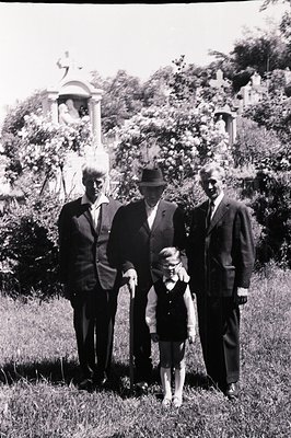 Formal portrait of three men and a young girl in a cemetery setting. Architectural details include a stone monument, likely a...