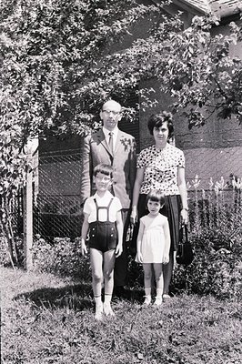 Family portrait, c. 1960s. A father in a suit, mother in a patterned dress, and two young children stand in front of a modest...