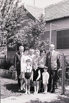 Family portrait, circa 1960s, shows three generations posed in front of a modest, tiled-roof home. Men wear suits & ties, wom...