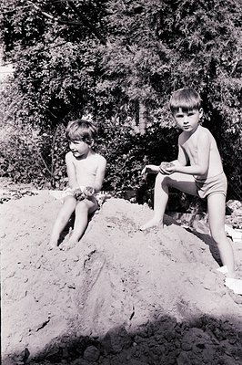 Two young boys sit atop a large sand dune. One is shirtless, holding handfuls of sand, while the other wears swim trunks and ...