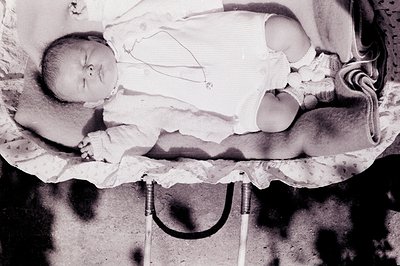 Black and white photo of a baby sleeping in a crib with decorative lace trim. The infant wears a simple white garment and a d...