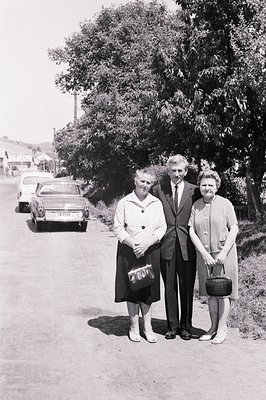 A posed black and white portrait depicts three formally dressed individuals standing on a rural road. A classic car is parked...