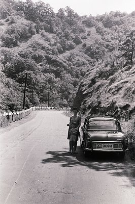 Monochrome image showing a woman and young child standing beside a vintage car on a winding mountain road. Dense, forested sl...