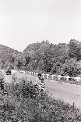 A woman holds a young child amidst roadside vegetation. Asphalt road bordered by a low stone wall leads toward a forested hil...