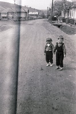 Two young boys, holding hands, walk down a dirt road lined with modest, older homes. Visible wear marks along the left edge s...
