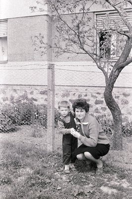 A young boy in overalls stands alongside his mother, who kneels near a wire fence. A stark, modernist building forms the back...