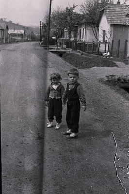 Two young boys stand on a rural road, dressed in traditional clothing - one in a patterned vest and dark trousers, the other ...