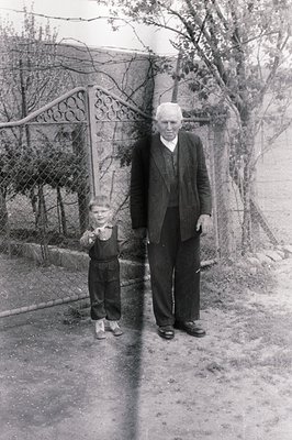 A young boy stands beside an older man, possibly a grandfather, outside a rustic home. The man wears a suit and tie; the boy,...