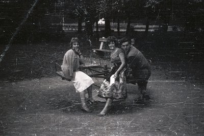 Three people seated on a picnic table in an outdoor setting. A woman in a dress, another in a skirt suit, and a man in a jack...