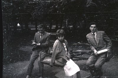 Three figures posed on a circular bench in a park setting. Two young men in suits flank a young woman, who appears pensive. O...