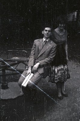 A formally attired man sits on a metal chair, holding sheet music. A woman in a floral dress stands beside him. Appears to be...