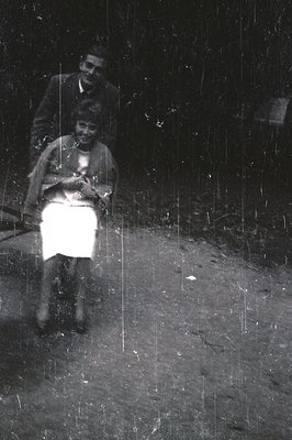 A seated woman in a tailored suit and a man in a jacket stand outdoors. The woman holds what appears to be a small book or pr...