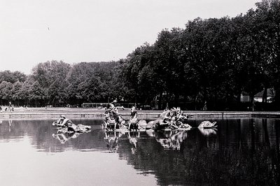 Ornate, bronze sculpture group depicting mythological figures in a fountain, reflected in still water. Formal landscaping & d...