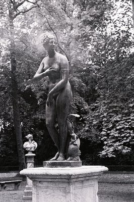 A bronze statue of a woman in a classical pose stands on a stone pedestal within a lush, leafy park setting. She appears to b...