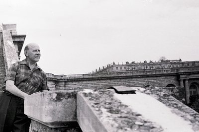 A man in a plaid shirt stands on a rooftop, leaning on a weathered stone parapet. Behind him stretches a monumental, ornate b...