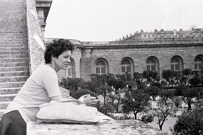 A woman in a simple top and skirt rests on a stone ledge, overlooking formal gardens and a large, ornate building with arched...