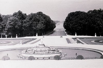 Formal French garden with tiered fountain, statues, and meticulously manicured hedges. View extends down a long, straight all...