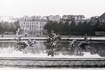 Ornate fountain sculptures reflecting in still water, set within a formal garden. Background reveals Parisian architecture—mu...