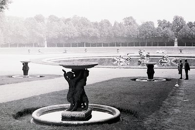 Formal garden scene with tiered fountains & classical statuary. Three fountains visible, central one depicts a figure support...