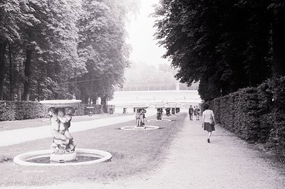 Ornate fountain and gravel path lead towards a formal garden facade. Several figures are visible in the distance, strolling a...