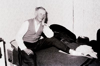 Man seated on a vintage sofa, showcasing a leg encased in a cast. He wears a waistcoat over a collared shirt and dark trouser...