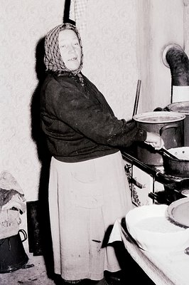 A woman in traditional clothing tends to a large stove in a rustic kitchen. She wears a dark dress, white apron, and headscar...