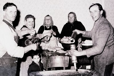 A family workshop scene, likely cheese or dairy production. Five figures are shown – a man in an apron, two women in headscar...