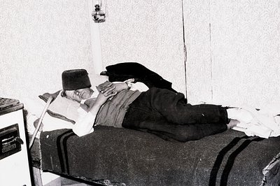Man lies on a narrow bed, propped up by pillows, wearing a traditional hat and vest. A medical device sits beside the bed in ...