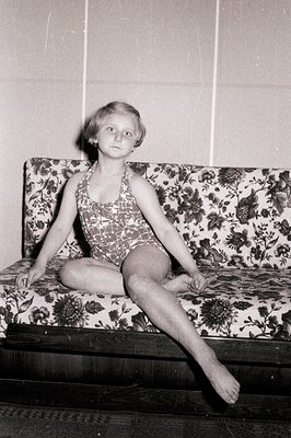 Young girl in a one-piece bathing suit, seated on a floral-patterned bench or sofa. Posed portrait, likely mid-20th century, ...