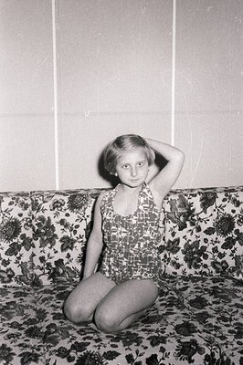 A young girl in a patterned one-piece swimsuit sits cross-legged on a floral-print sofa. She's touching her hair, facing the ...