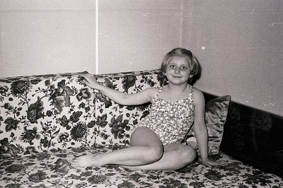 A young girl, circa 1960s-70s, sits cross-legged on a floral-patterned couch, wearing a one-piece bathing suit. Her arm is ex...