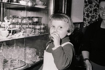 A young child stands before a glass display case filled with delicate china and figurines, appearing to savor a treat. The ch...