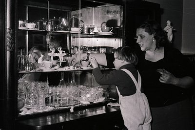 Young boy in overalls examines a glass cabinet filled with china, silver, and figurines. A woman stands behind him, her expre...