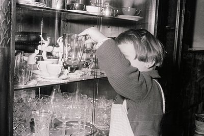 A young boy reaches for a glass goblet within a crowded china cabinet. The cabinet displays delicate glassware, ornate figuri...
