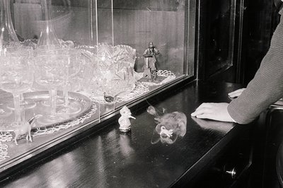 Black and white photograph shows a glass display case with decorative figurines and cut-glassware. The scene includes a man i...