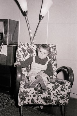 Young boy sits in a patterned armchair, wearing overalls and a dark sweater. A vintage floor lamp looms behind. Mid-century m...