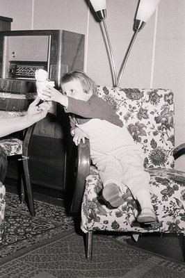 A young child, clad in a red sweater and patterned trousers, sits in a patterned armchair, reaching for an object held by an ...