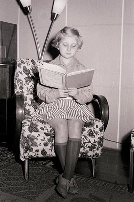 Young girl engrossed in a book while seated in a floral-patterned armchair. The decor suggests a mid-century interior with a ...