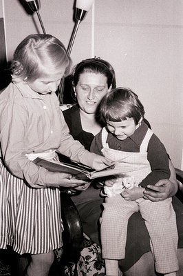 A mother sits in a patterned armchair, reading a book to two young children. Both children appear engaged, one standing and p...