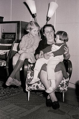 A mother cradles a baby in a patterned armchair, while a young girl sits nearby. Mid-century modern decor, including a radio ...