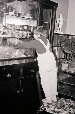 A young child in overalls stands on a patterned chair, reaching towards a tall, ornate glass display cabinet. The cabinet hol...